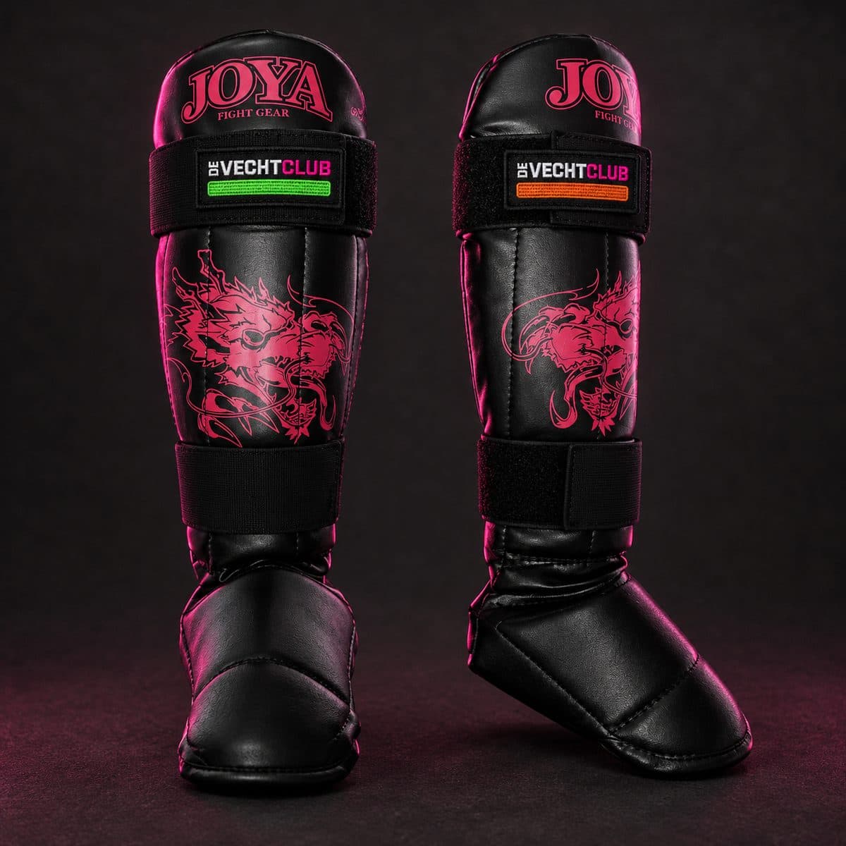Two pink and black kids' shin guards with embroidered De Vechtclub tags featuring a green and an orange stripe