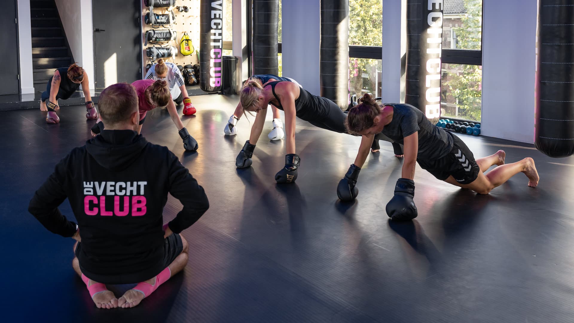 Een trainer in een zwarte De Vechtclub-hoodie zit op de mat en kijkt toe terwijl sporters met bokshandschoenen aan opdrukken.