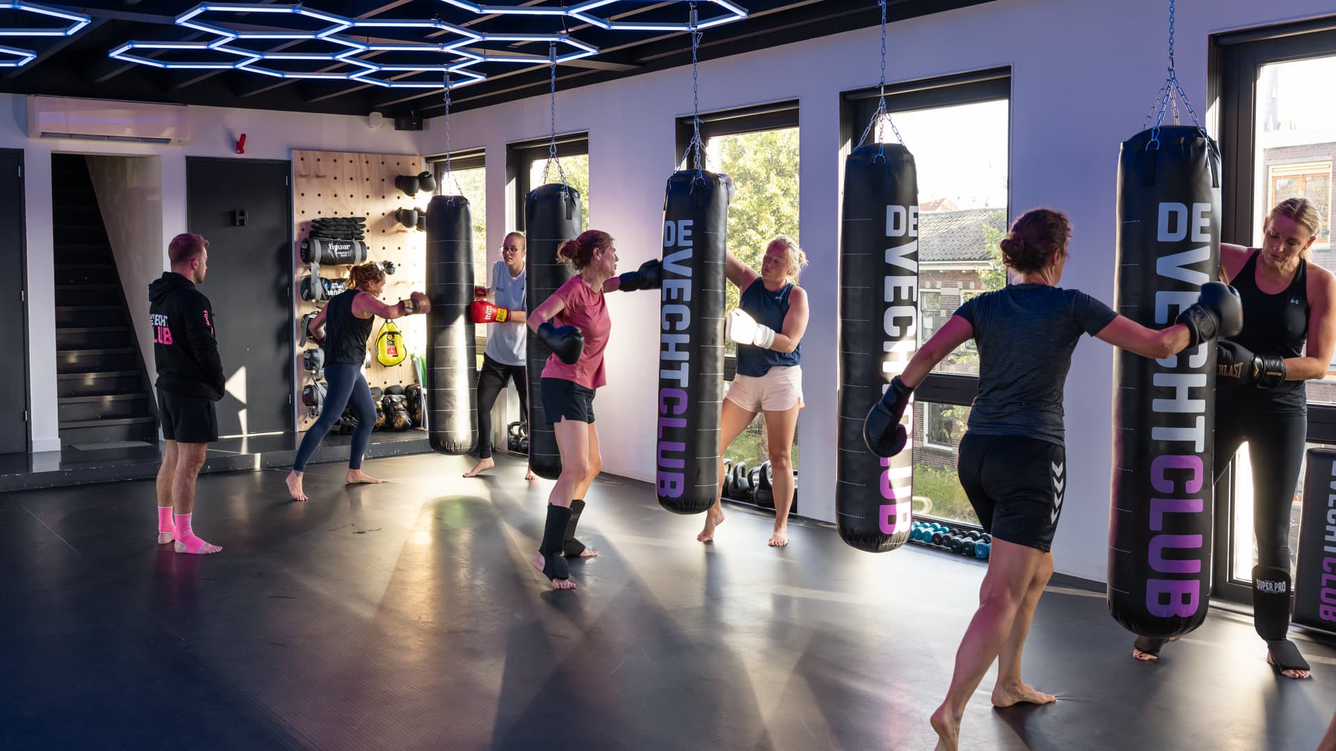 Zaktraining in de zaal aan de Achtergracht: sporters stoten en trappen op zwarte bokszakken met het De Vechtclub-logo, een trainer in een zwarte clubhoodie kijkt toe onder de zeshoekige plafondverlichting.
